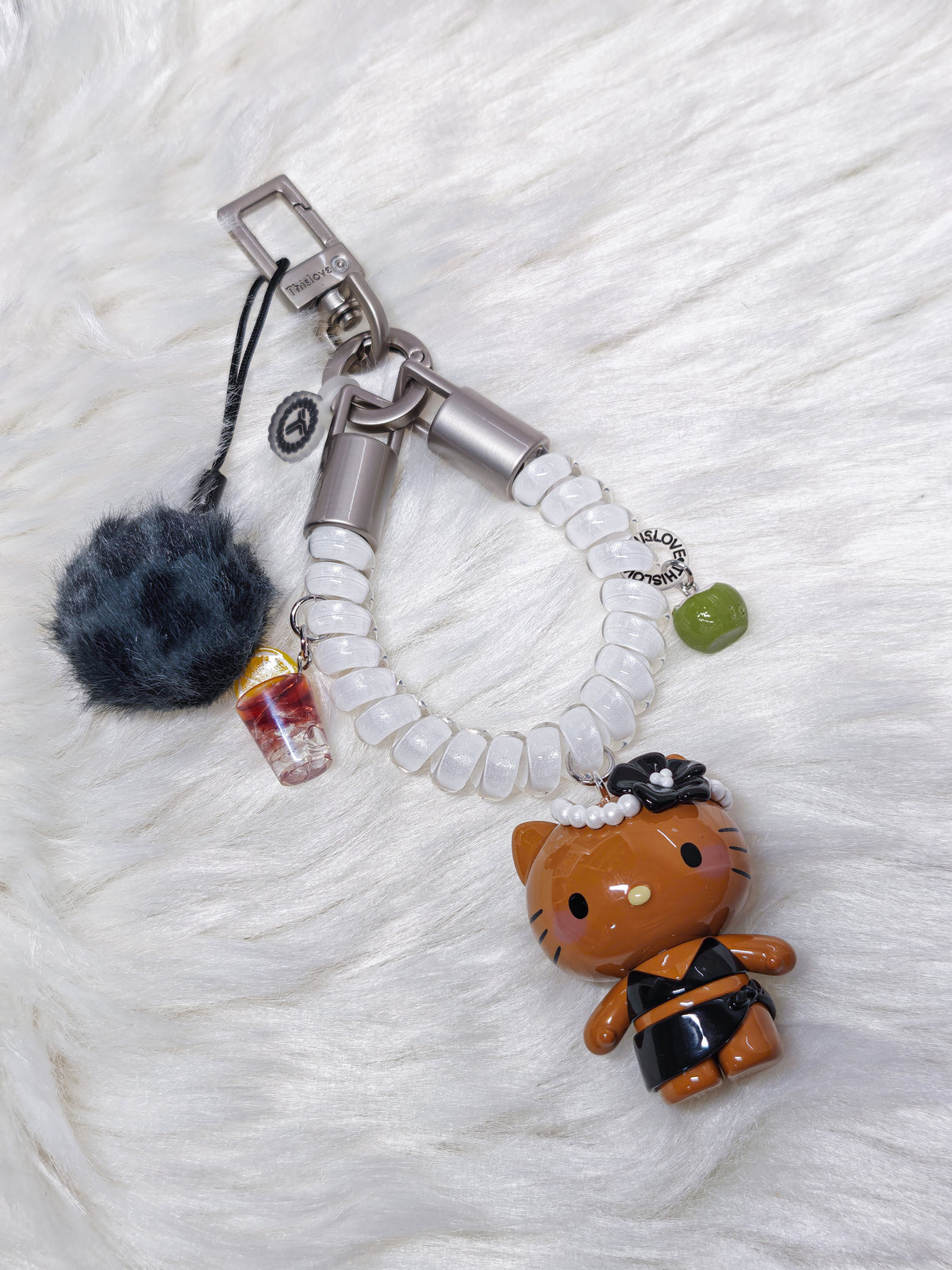A Hello Kitty themed keychain bag charm set featuring a brown Hello Kitty figure in a black outfit, a white beaded strap, a dark gray pom-pom, a miniature drink charm, and a small green bead, displayed on a white faux fur background.