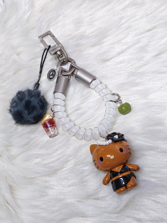 A Hello Kitty themed keychain bag charm set featuring a brown Hello Kitty figure in a black outfit, a white beaded strap, a dark gray pom-pom, a miniature drink charm, and a small green bead, displayed on a white faux fur background.