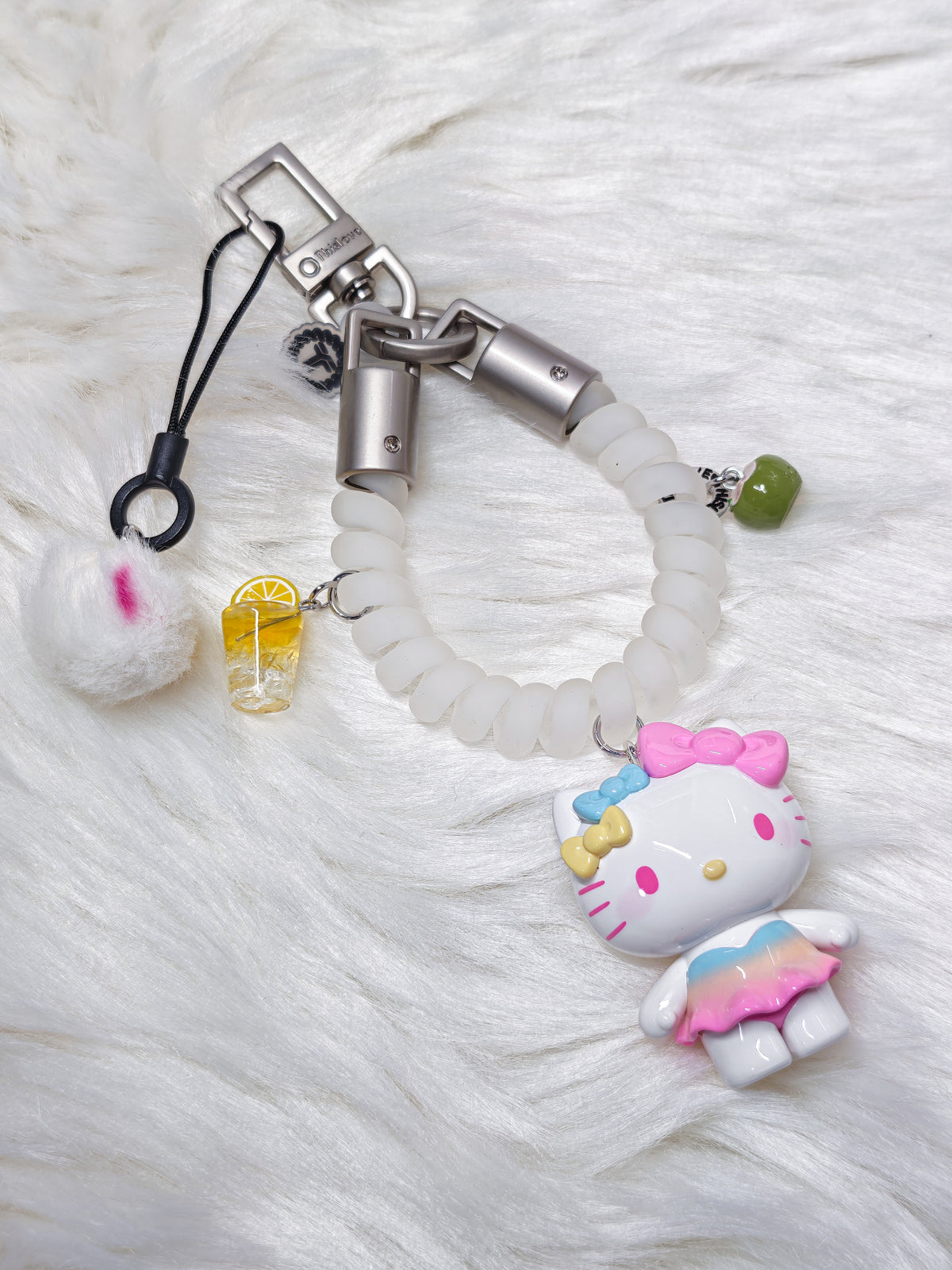 A Hello Kitty themed phone strap keychain featuring a white coiled cord, a silver metal clasp, a white Hello Kitty charm with a pink bow and pastel skirt, a small yellow drink charm, and a white pom-pom accessory, all displayed on a white faux fur background.